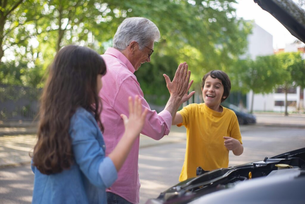 Cheerful grandfather and grandchildren high-five near car outdoors, enjoying a sunny day together.